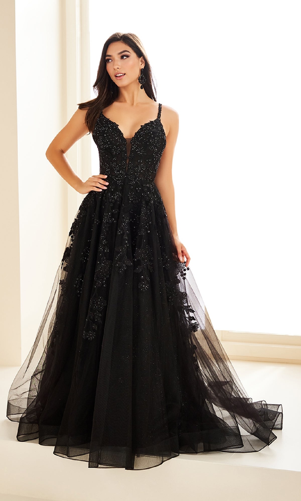 A woman stands indoors wearing the Ellie Wilde EW36015 long prom dress, featuring a fitted corset bodice with thin straps and a full black glitter tulle skirt adorned with sparkling details and floral accents.